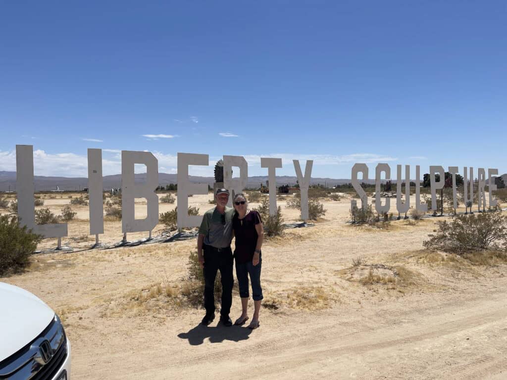Art & Julie of The Places Where We Go at Liberty Sculpture Park in Yermo, CA - standing in front of Liberty Sculpture Park signage
