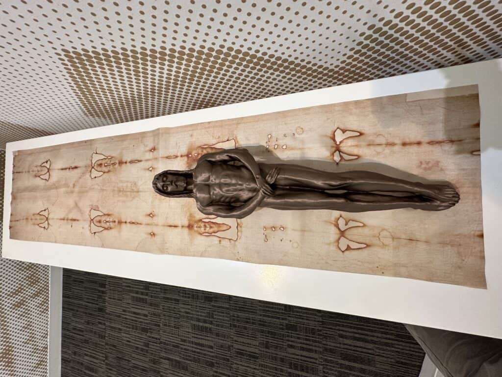 Full-sized Shroud of Turin reproduction displayed on an illuminated wall with a reclining sculpture of Christ in the exhibition gallery.